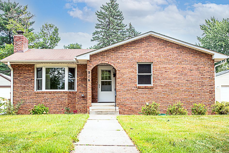 A single-story brick house features a welcoming front entrance with three steps leading to the door. A neat lawn surrounds the home, and large windows allow natural light to fill the interior. Green shrubs are planted along the foundation, adding natural beauty.