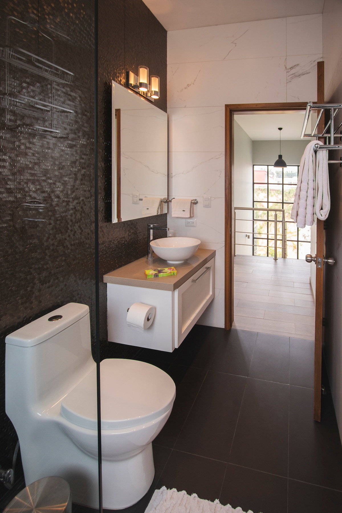 A modern bathroom features sleek black tiles and a stylish sink mounted on a wooden vanity. A well-lit mirror is above the sink, complemented by two minimalist wall sconces. A neatly placed towel rack and an adjacent frosted-glass door provide access to a well-lit area beyond.