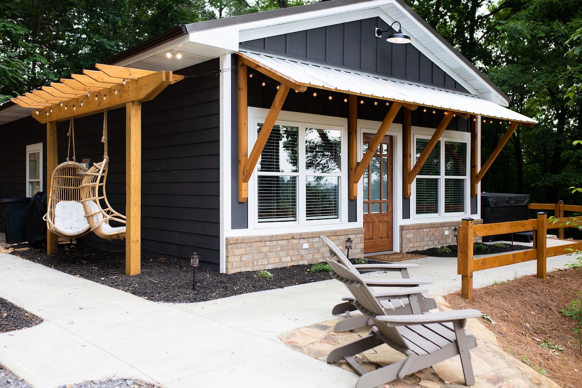 The exterior of the cottage features a welcoming porch with a hanging chair and decorative string lights. A pathway leads to the entry door, bordered by rustic wooden fences and seating areas. Large front windows allow natural light to fill the interior.