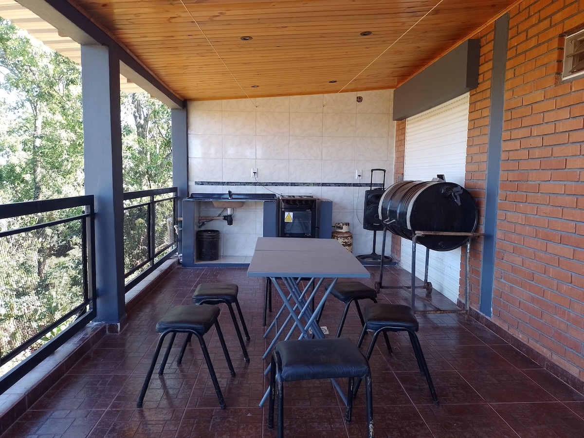 An outdoor terrace is presented with a spacious table surrounded by six black chairs. A built-in kitchen area features a countertop and a grill, set against a backdrop of natural greenery visible through the railing.