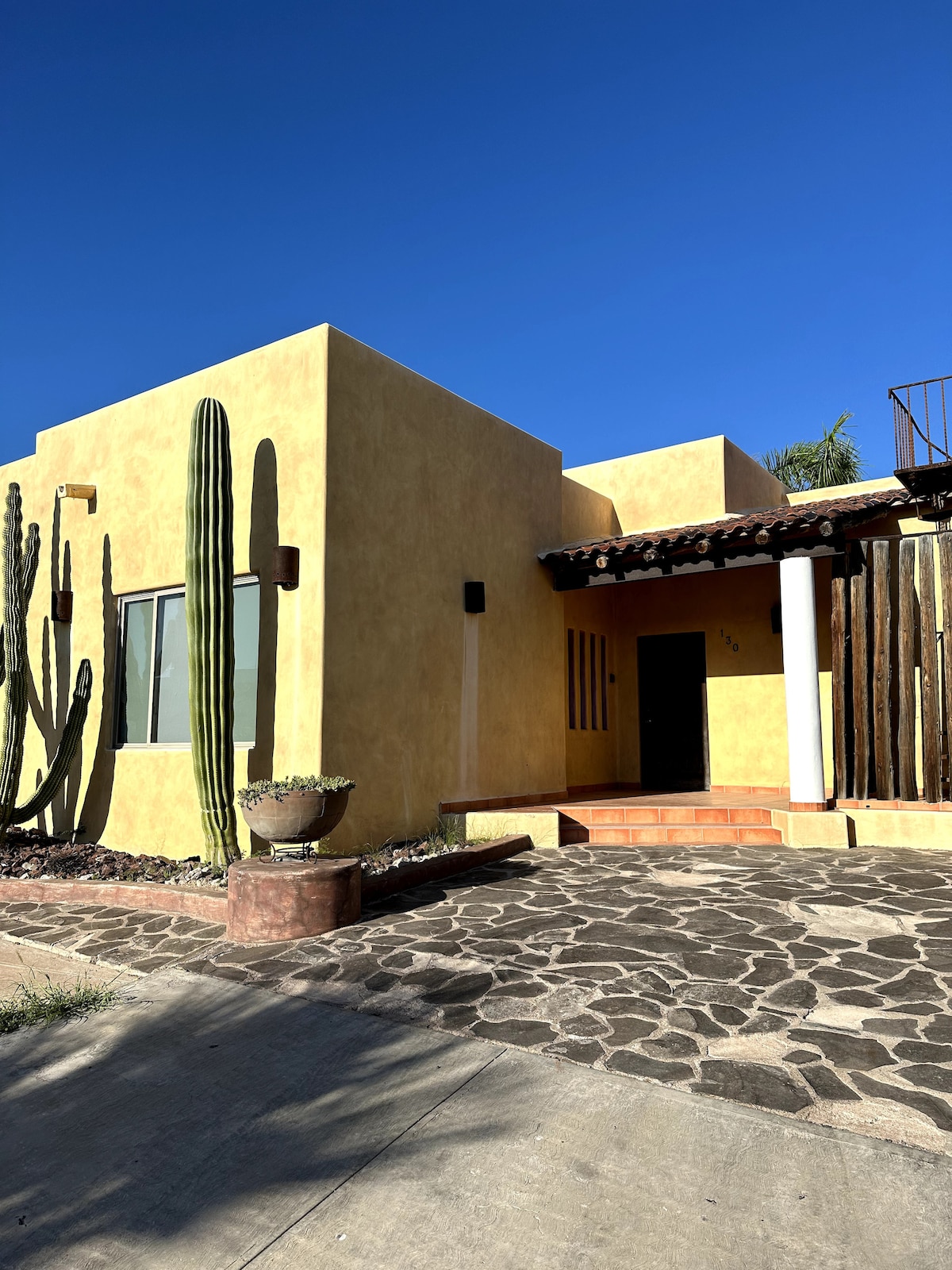 The exterior of a vibrant yellow house is presented, featuring tall green cacti and a stone pathway leading to the entrance. The structure showcases a mix of modern and traditional architectural elements with wooden accents and large windows under a clear blue sky.