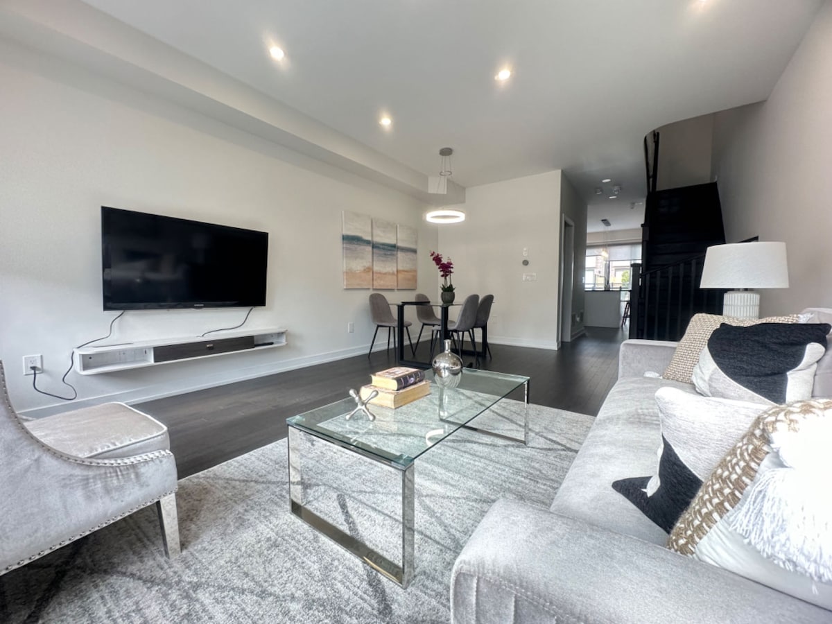 A modern living area features a flat-screen television on the wall opposite a plush sofa and two stylish chairs. A glass coffee table sits at the center, while a dining table with four chairs is positioned nearby. Bright overhead lighting complements the open layout.