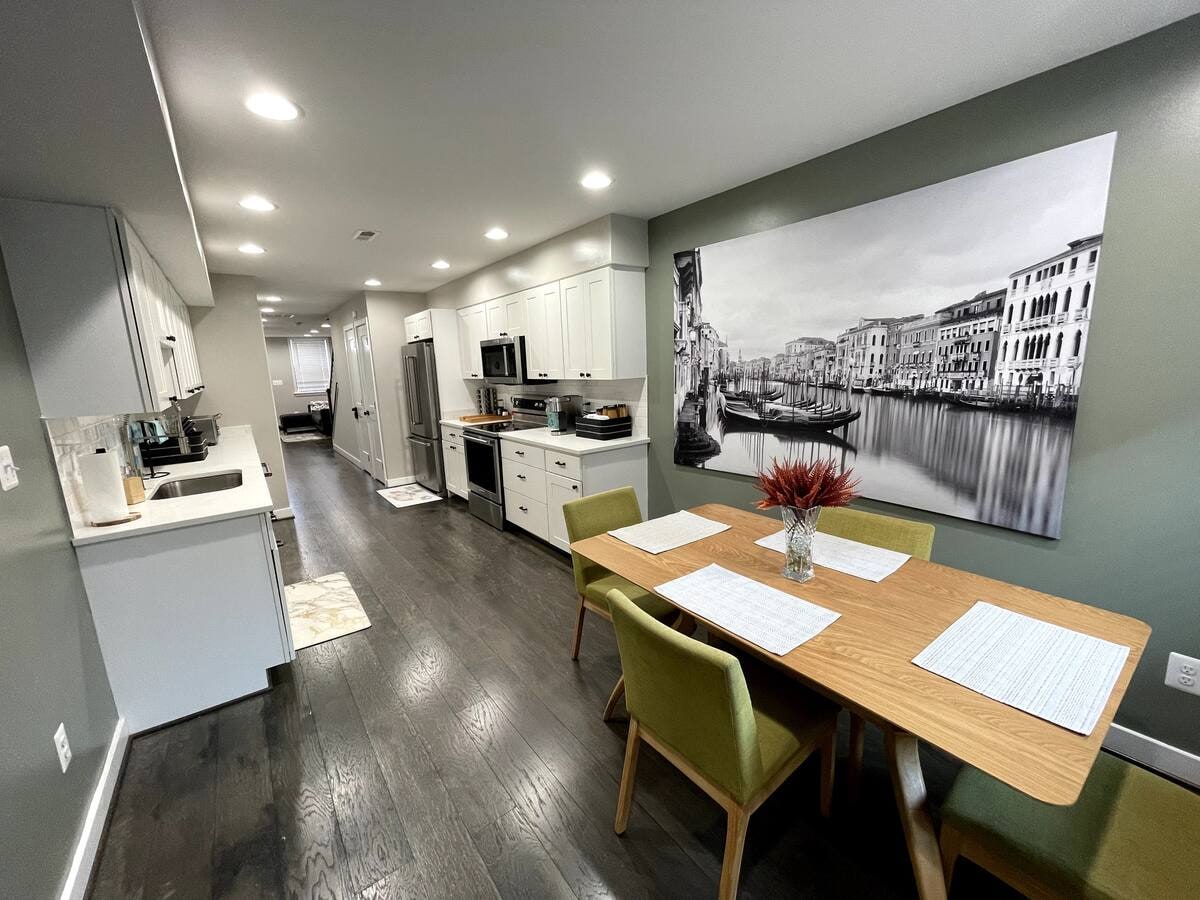 A modern kitchen and dining area feature light cabinetry and stainless steel appliances. A dining table with green chairs sits in the foreground, complemented by a large black and white wall mural depicting a scenic waterfront. Soft lighting enhances the room's spacious feel.