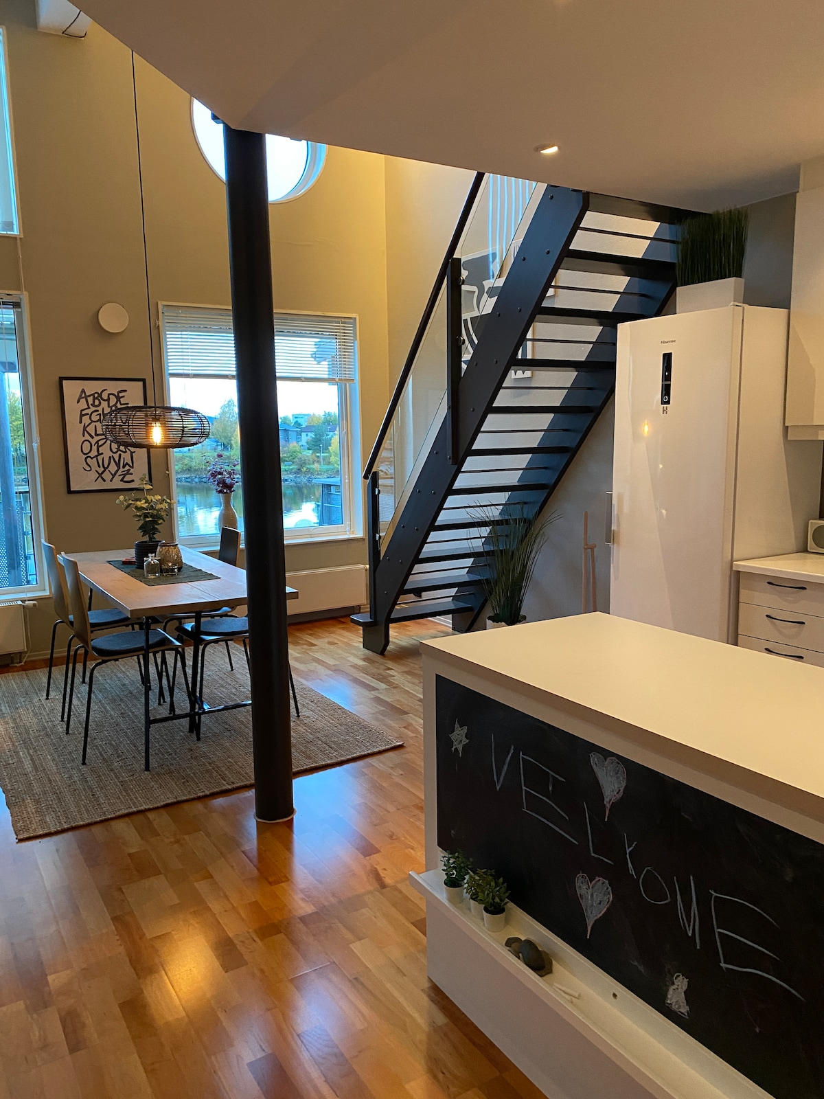 An open-plan living space features a modern staircase leading to the upper floor. Large windows admit natural light, illuminating a dining area with a wooden table and charming pendant light. A kitchen is visible, distinguished by sleek white cabinetry and a chalkboard welcome sign.