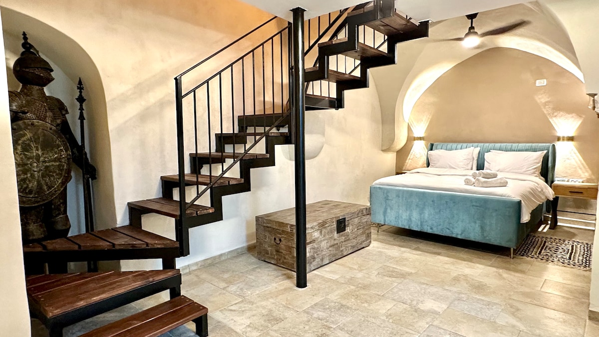 A multi-level living space features a spiral staircase with dark wooden steps leading to an upper area. A spacious bed with light linens is positioned against a muted wall, while a rustic storage chest and decorative elements enhance the ambiance.