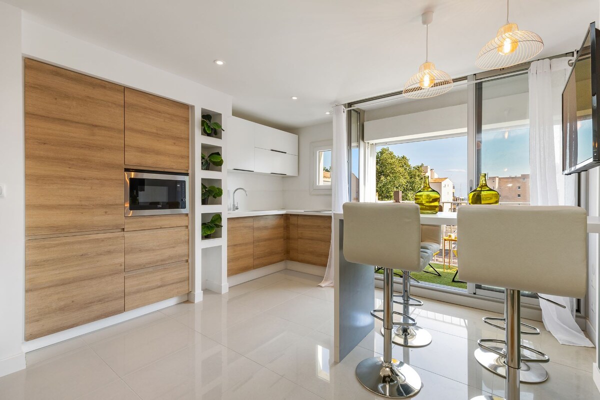 A modern kitchen is presented with sleek cabinetry and a minimalistic design. A dining area is featured with two high-backed stools, complemented by a view of the outdoors through expansive glass doors. Natural light brightens the space, enhancing the clean aesthetics.