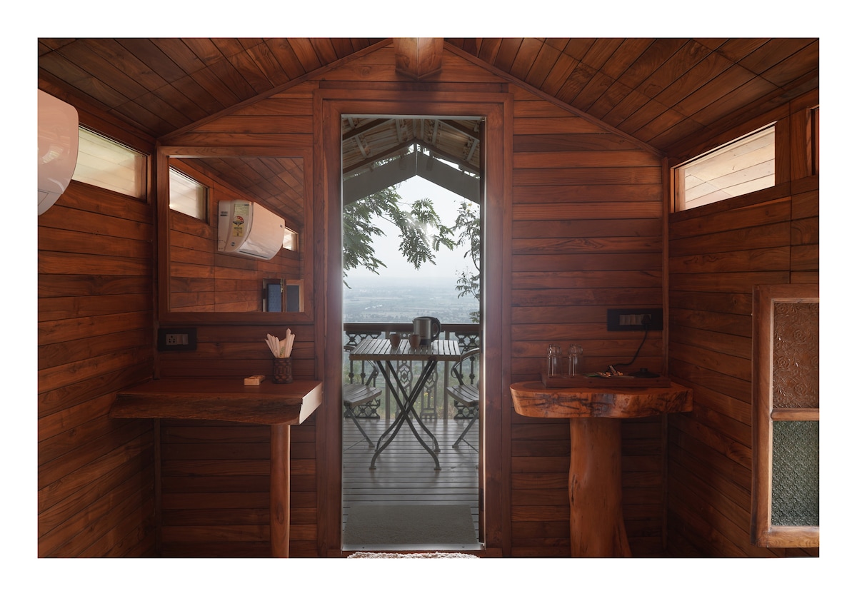 A wooden interior showcases natural textures and warm tones. A small table and a wooden counter are visible, complemented by a bowl and glasses. The open door leads to a balcony with a scenic view of the mountains and valley.
