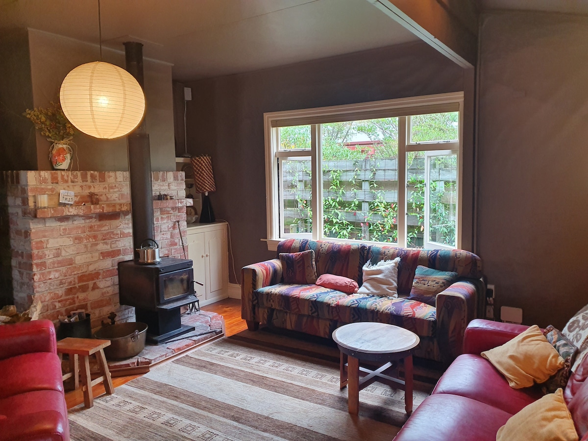 A cozy living area is featured with a colorful patterned sofa and a red leather couch, providing ample seating. A wood-burning stove sits on a brick hearth, while a round wooden table is positioned in front. Natural light enters through a large window overlooking a garden.