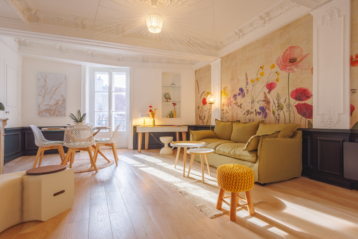 A bright living space is characterized by floral wall art and natural light streaming through large windows. A comfortable sofa is complemented by a small accent stool and a minimalist coffee table, while a dining area is thoughtfully arranged with modern seating.