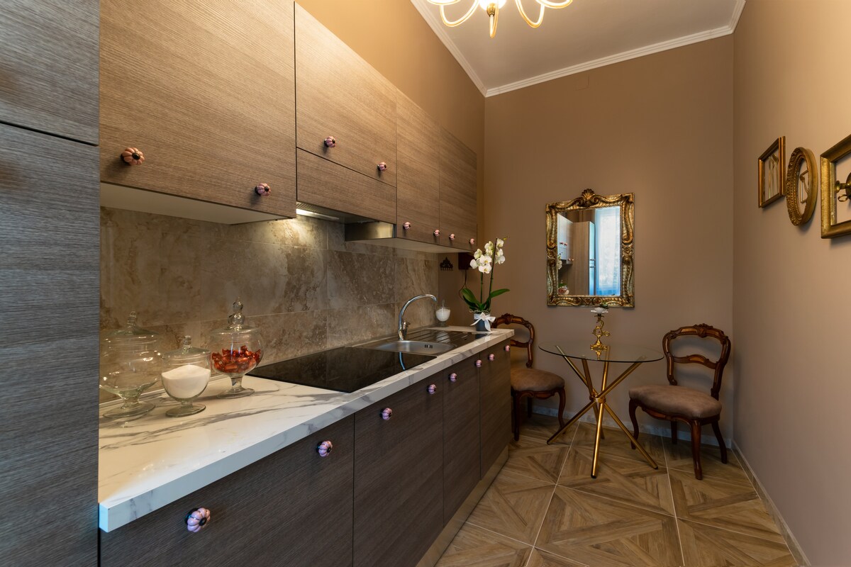 The kitchen area features modern cabinetry with a rich wood finish, complemented by a marble countertop. A small round table with two chairs is positioned near a mirror adorned with a decorative frame. Natural light highlights a plant and stylish decor.