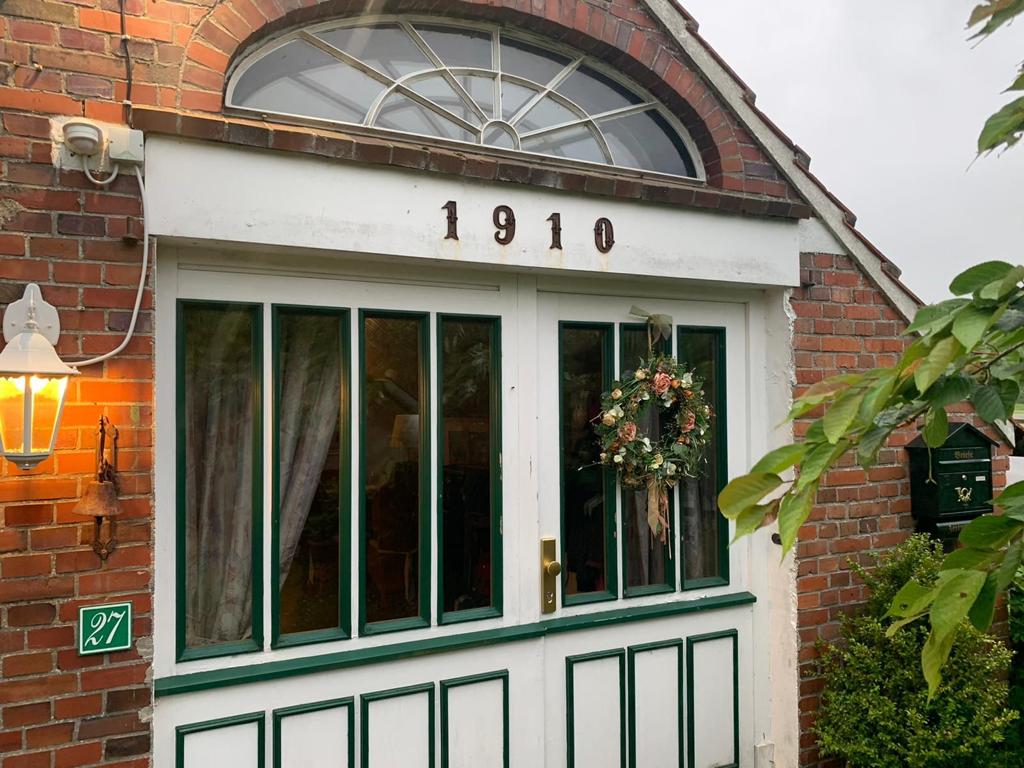 The entrance features a classic design with a white door framed by green trim. Above the door, the year '1910' is prominently displayed. A floral wreath adorns the door, while a brick facade and subtle greenery create a welcoming atmosphere.