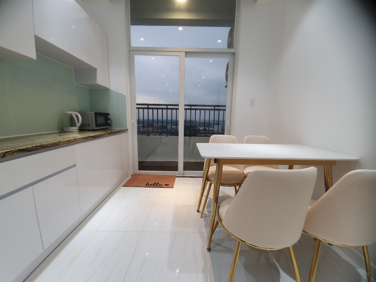 A dining area is illustrated with a white table surrounded by four light-colored chairs. A panoramic glass door provides views of the balcony and cityscape beyond. Kitchen appliances, including a kettle and microwave, are positioned on the counter, enhancing the modern design.
