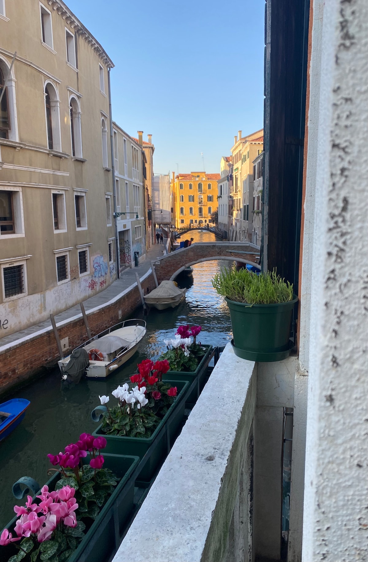 A view is presented from a balcony overlooking a narrow canal lined with traditional Venetian buildings. Several boats are anchored along the waterway. Flower pots with blooming colorful flowers enhance the scene, adding a touch of greenery and vibrancy to the historic surroundings.