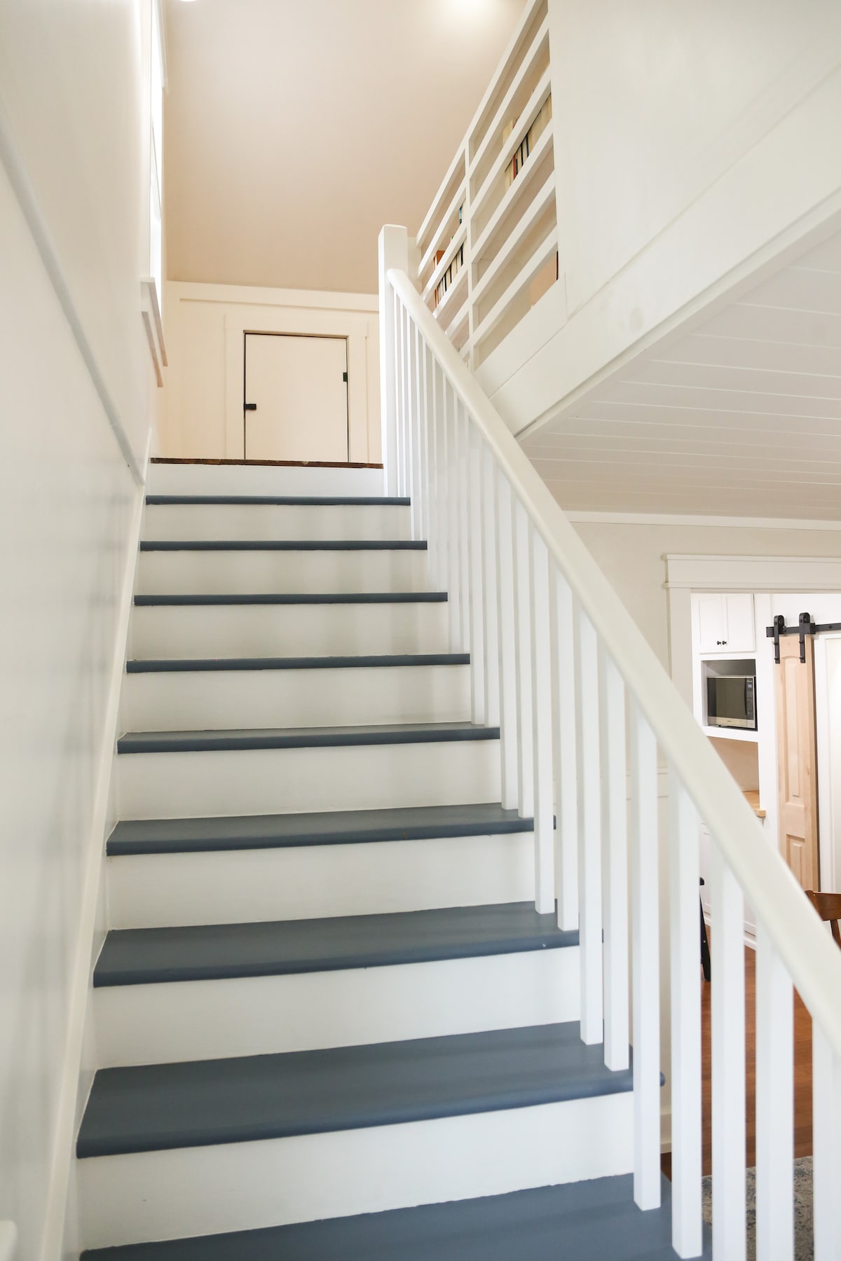 A set of light-colored stairs with a soft blue finish leads upward, supported by white handrails. The upstairs landing is visible, featuring a small door at the top. Well-lit surroundings enhance the clean and inviting aesthetic of the space.