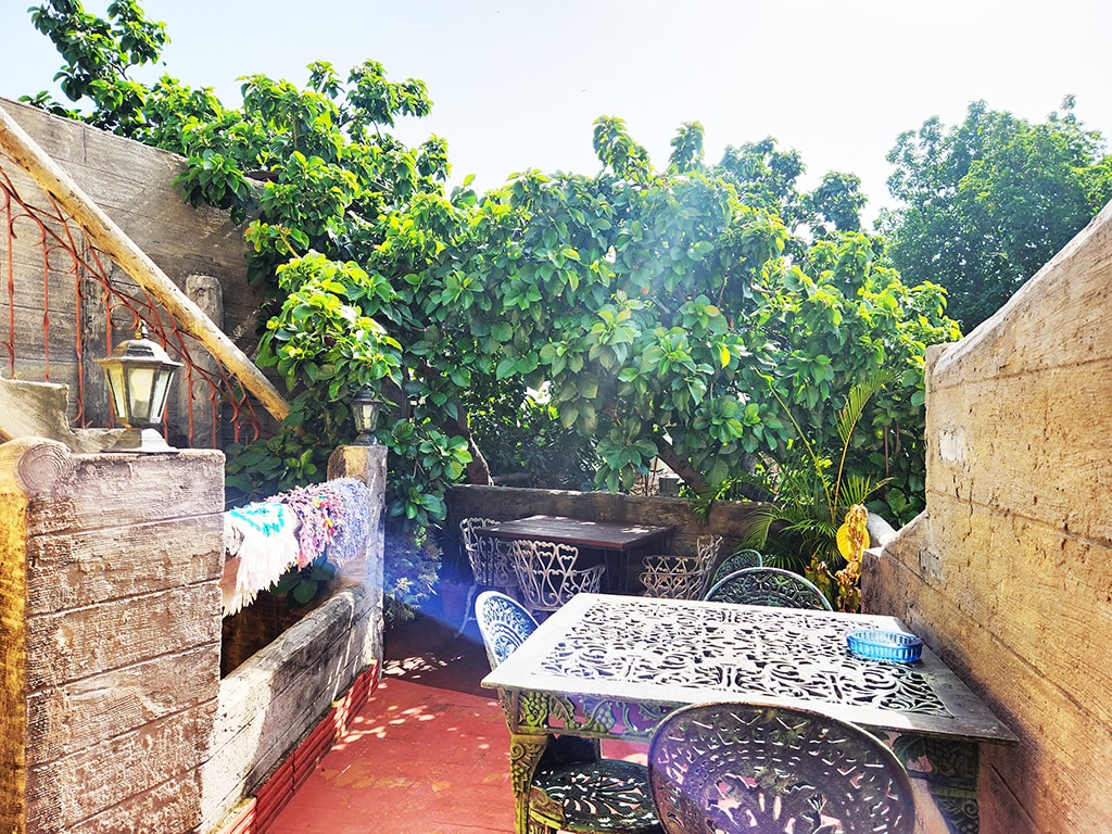 A rooftop terrace is surrounded by lush greenery, creating a serene outdoor space. A dark metal table is accompanied by matching chairs, inviting guests to relax. Warm sunlight brightens the area, highlighting its tranquil setting and connection to nature.