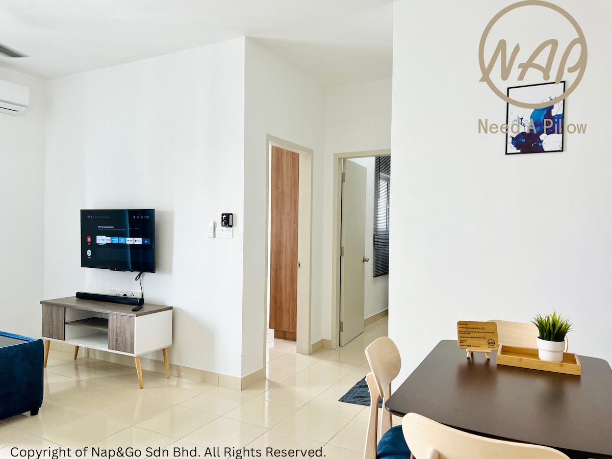 A bright living space features a wall-mounted television, a dining table, and a small kitchenette area. Air conditioning is visible on the wall, and a door leads to a bathroom. Natural light fills the area, which is furnished with neutral tones.