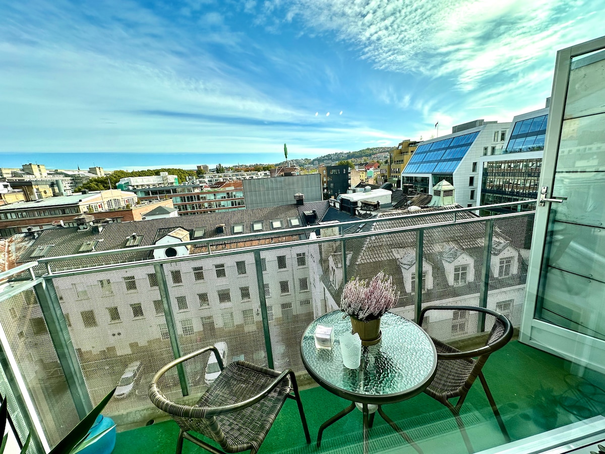 A balcony is seen with a glass-topped table and two woven chairs, overlooking the urban landscape. The sky is bright with scattered clouds, and rooftops of nearby buildings are visible in the background.