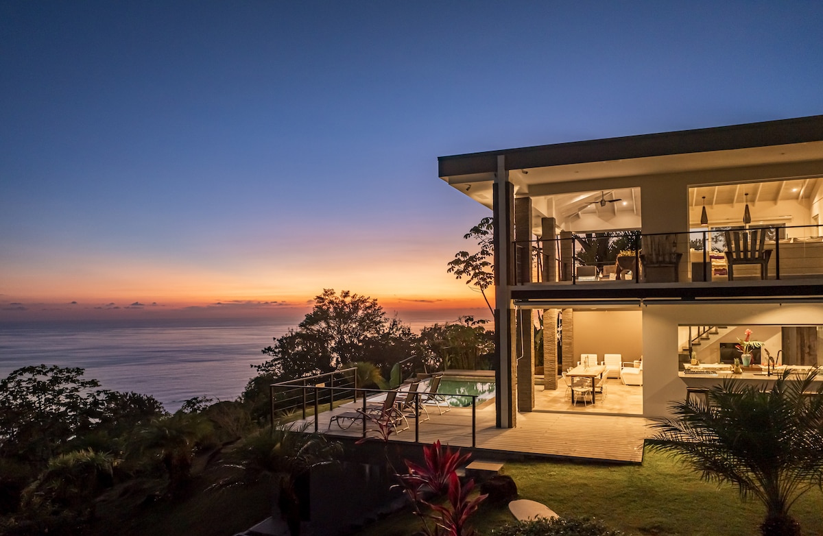 A modern two-story home is framed against a vibrant sunset sky. The structure features large glass doors showcasing the indoor seating area, while a private pool is visible outdoors among lush greenery. The tranquil ocean serves as an expansive backdrop.