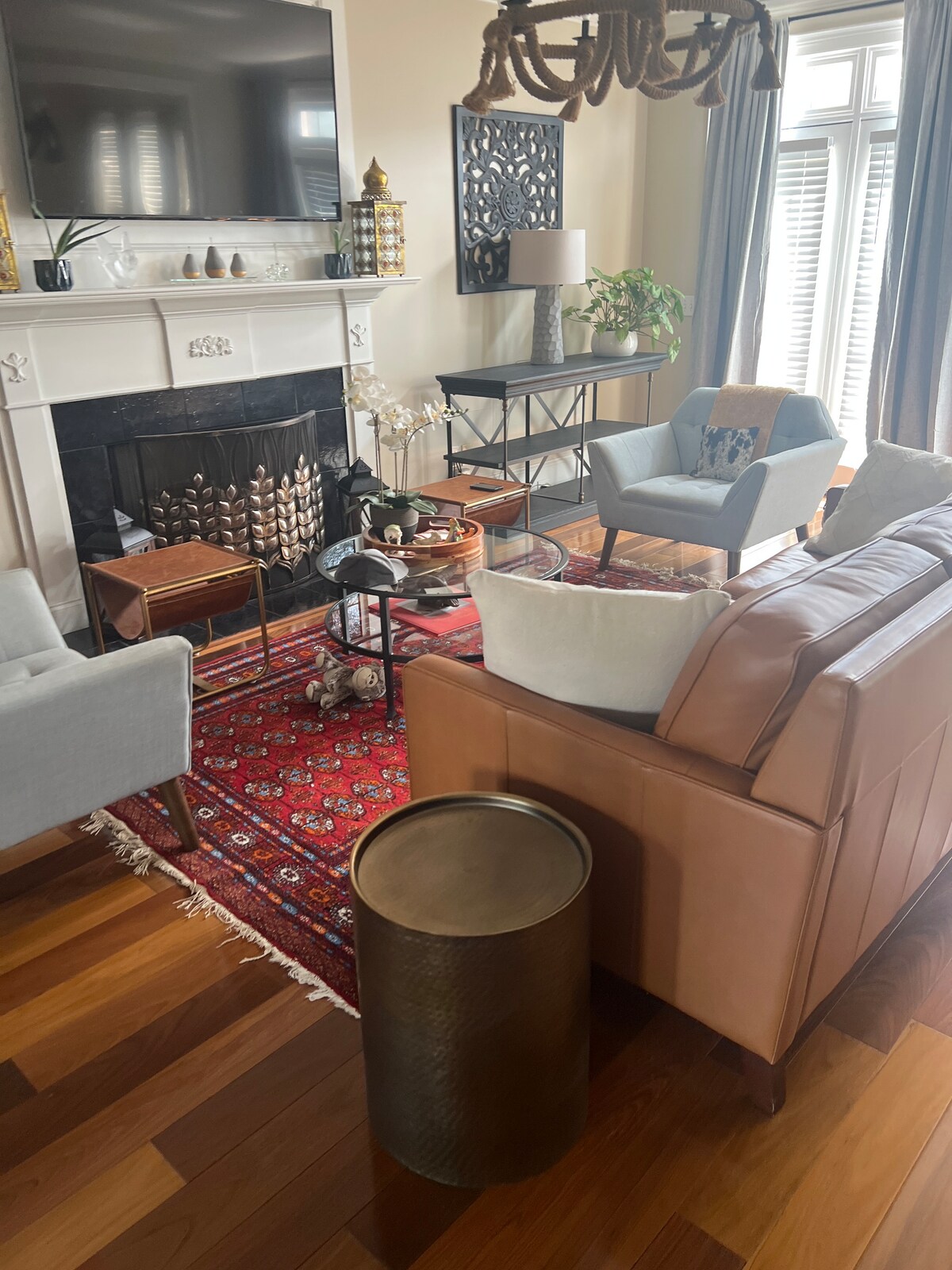 A well-furnished living area features a mixture of seating options, including light-colored armchairs and a brown leather sofa. A patterned area rug anchors the space, and a coffee table is surrounded by decorative elements. Bright light filters through sheer window treatments.