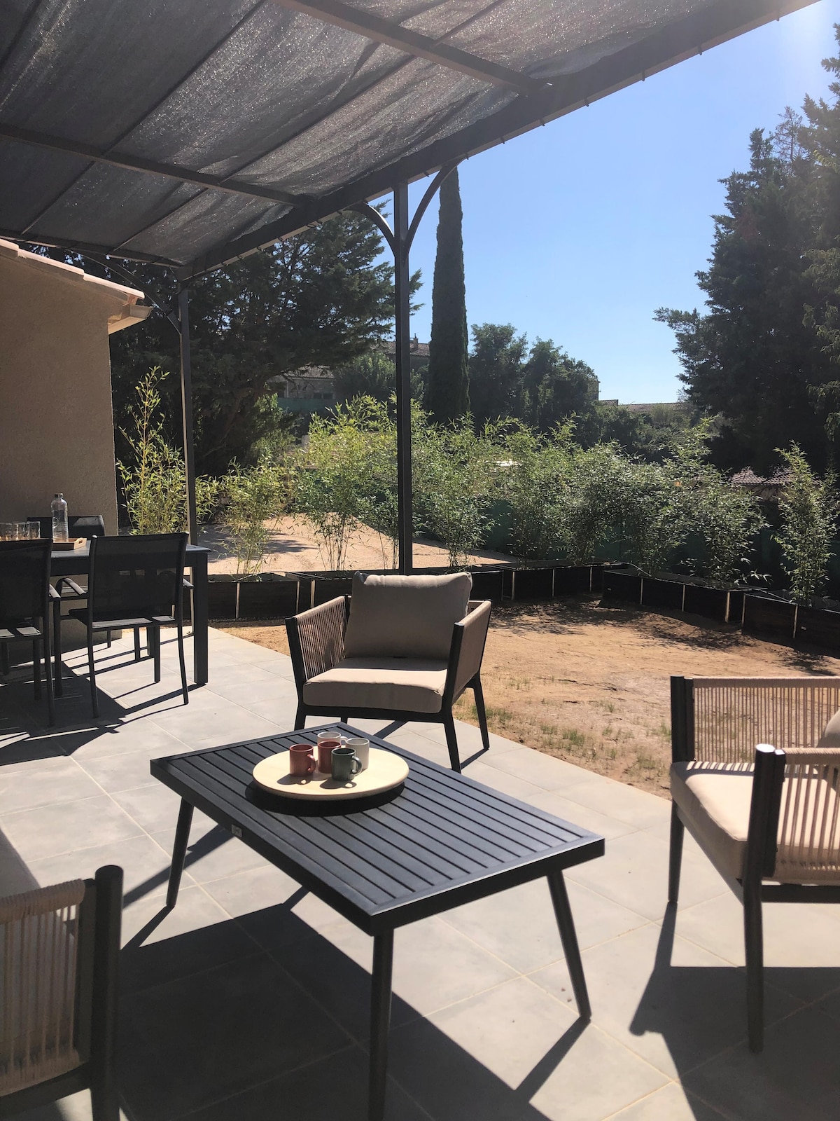 An outdoor seating area features a black table and four chairs, shaded by a canopy. Green shrubs are visible in the background, alongside a light-colored home. The space is designed for relaxation with a calm, structured layout.