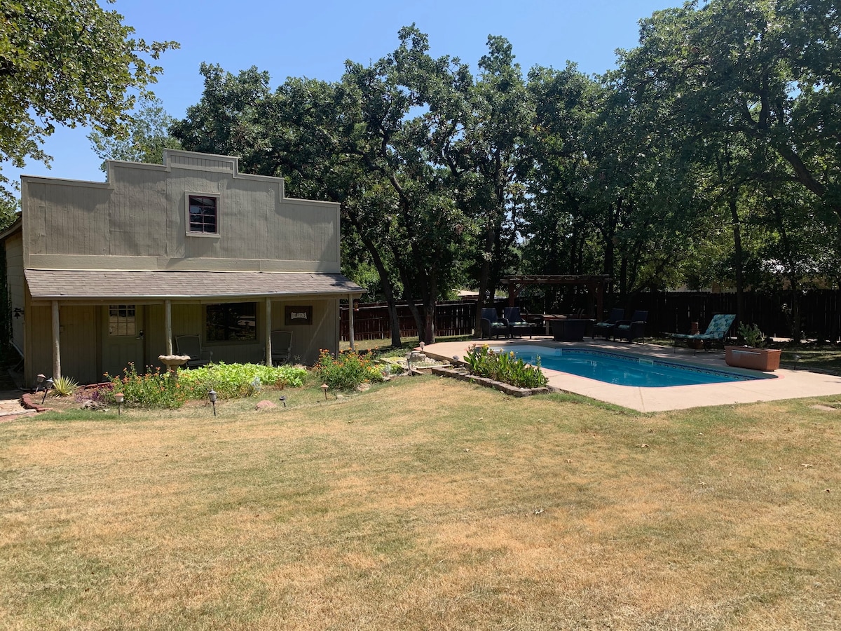 A spacious outdoor area is highlighted by a neatly maintained lawn, surrounded by trees for shade. The two-story guesthouse is situated nearby, with a covered patio visible. A swimming pool is located to one side, accompanied by lounge chairs and a stone path leading to the well-kept garden.