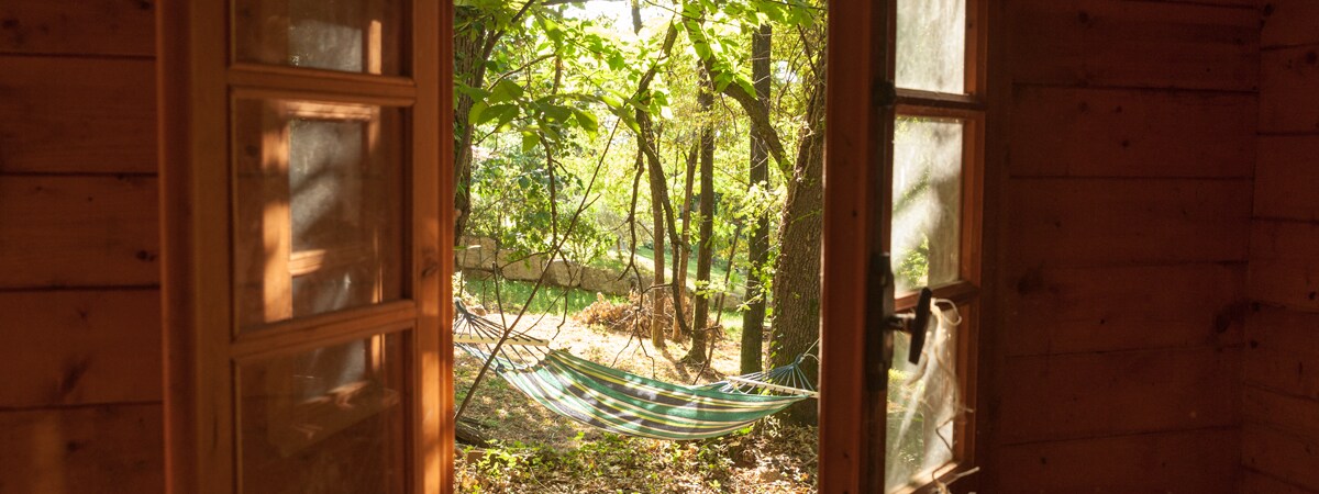 Double doors open to a serene outdoor scene, framed by sunlight filtering through trees. A colorful hammock is visible, suspended between two trees in a peaceful wooded area, inviting relaxation and respite.