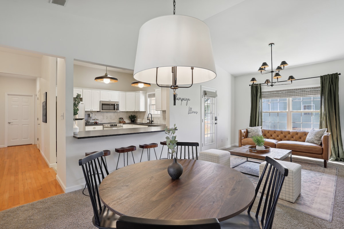 A spacious dining area features a round wooden table surrounded by dark wooden chairs, illuminated by a large pendant light. The open layout connects to a modern kitchen with stainless steel appliances and a bright living room, showcasing a comfortable sofa and elegant decor.