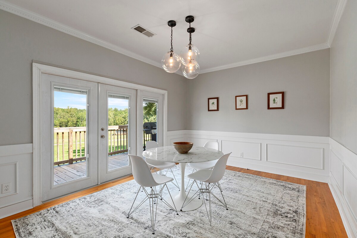 A dining area features a round marble table surrounded by white chairs. Soft lighting is provided by decorative pendant fixtures above. Double glass doors lead to a deck, framing a view of the green pasture beyond. Neutral colored walls and wainscoting complete the serene space.