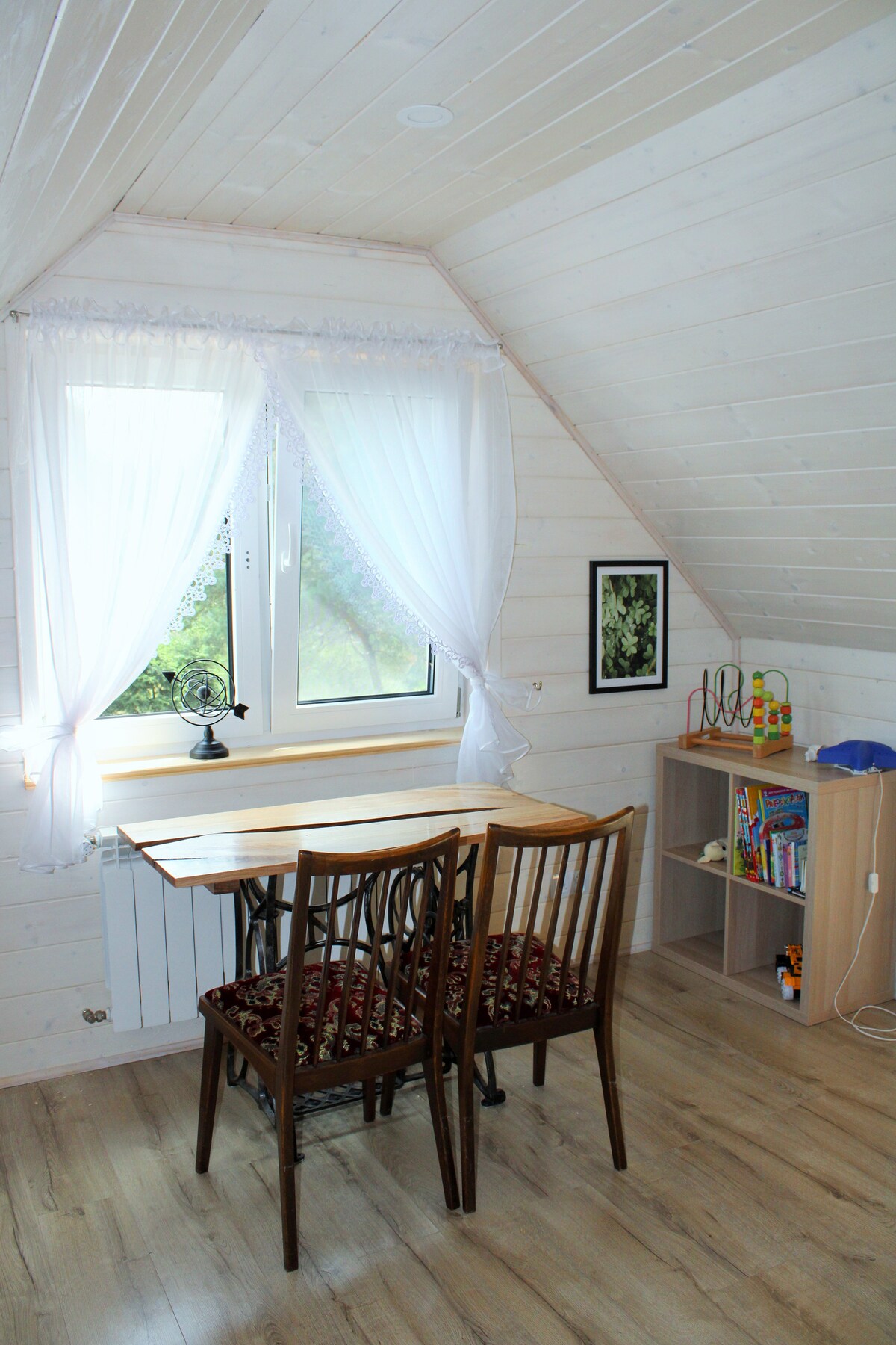A light and airy corner of the room features a wooden table with two chairs beneath a window adorned with sheer white curtains. Natural light filters in, illuminating a small bookshelf nearby filled with various items and a framed piece of art on the wall.