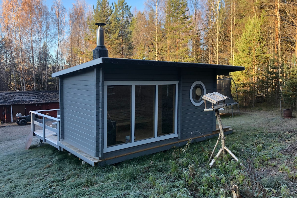 A modern, single-story blue-gray cabin is set within a forested area. Large glass windows provide views of the surrounding nature. A circular window adds character to the facade. A wooden deck extends outside, accompanied by an artistic bird feeder.