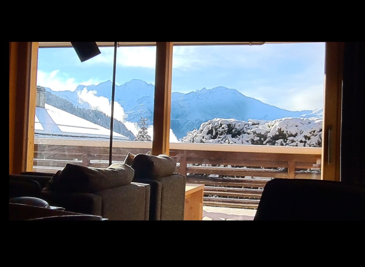 A view from the apartment showcases snowy mountains under a clear blue sky. Large windows provide ample natural light, framing the serene landscape and highlighting the outdoor balcony space. Cozy seating is positioned nearby, inviting relaxation while enjoying the vistas.
