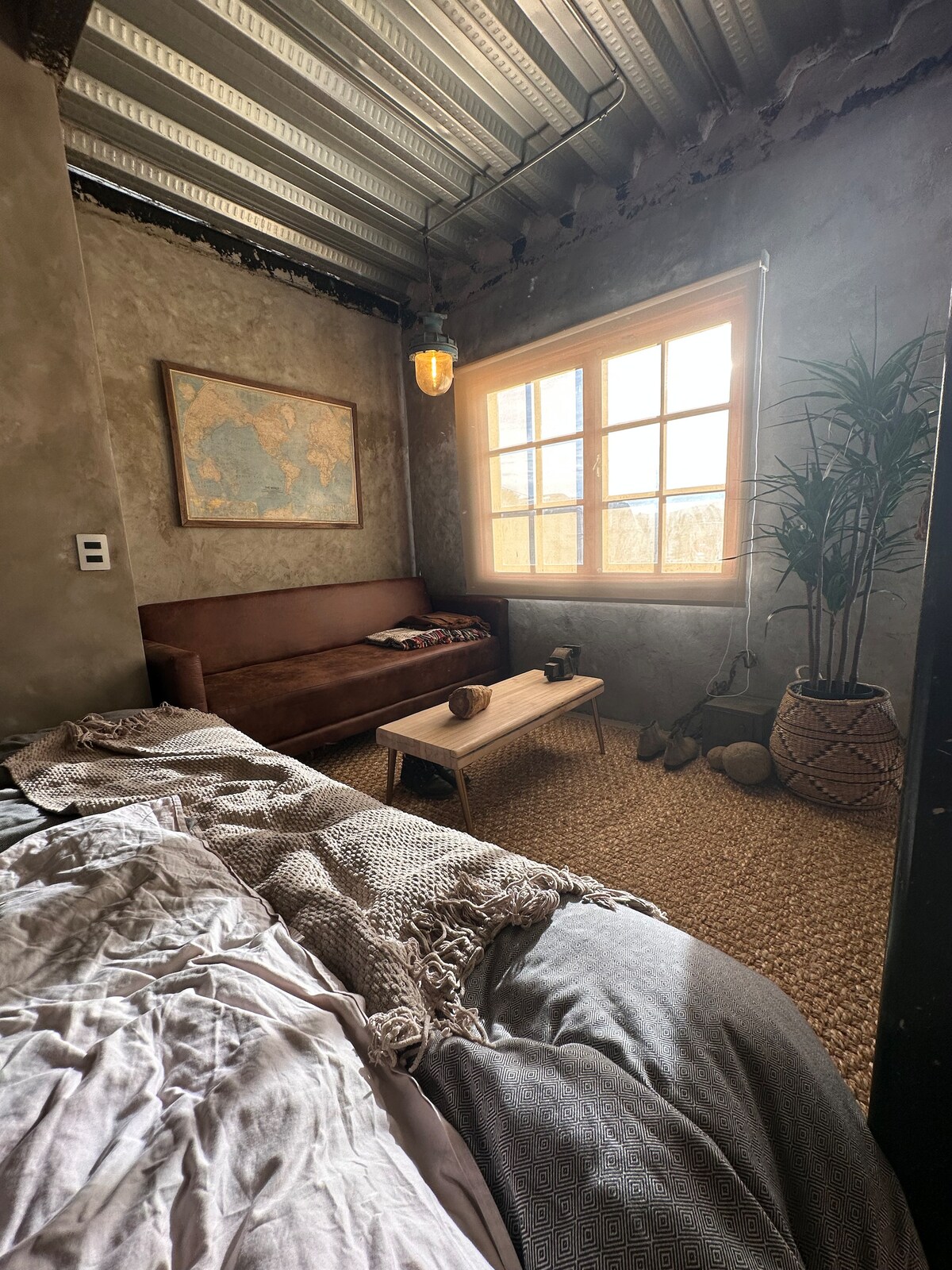 A rustic industrial studio features a cozy sitting area with a brown sofa and a small coffee table. A large window allows natural light to illuminate the space, while a potted plant and a world map artwork enhance the inviting atmosphere.