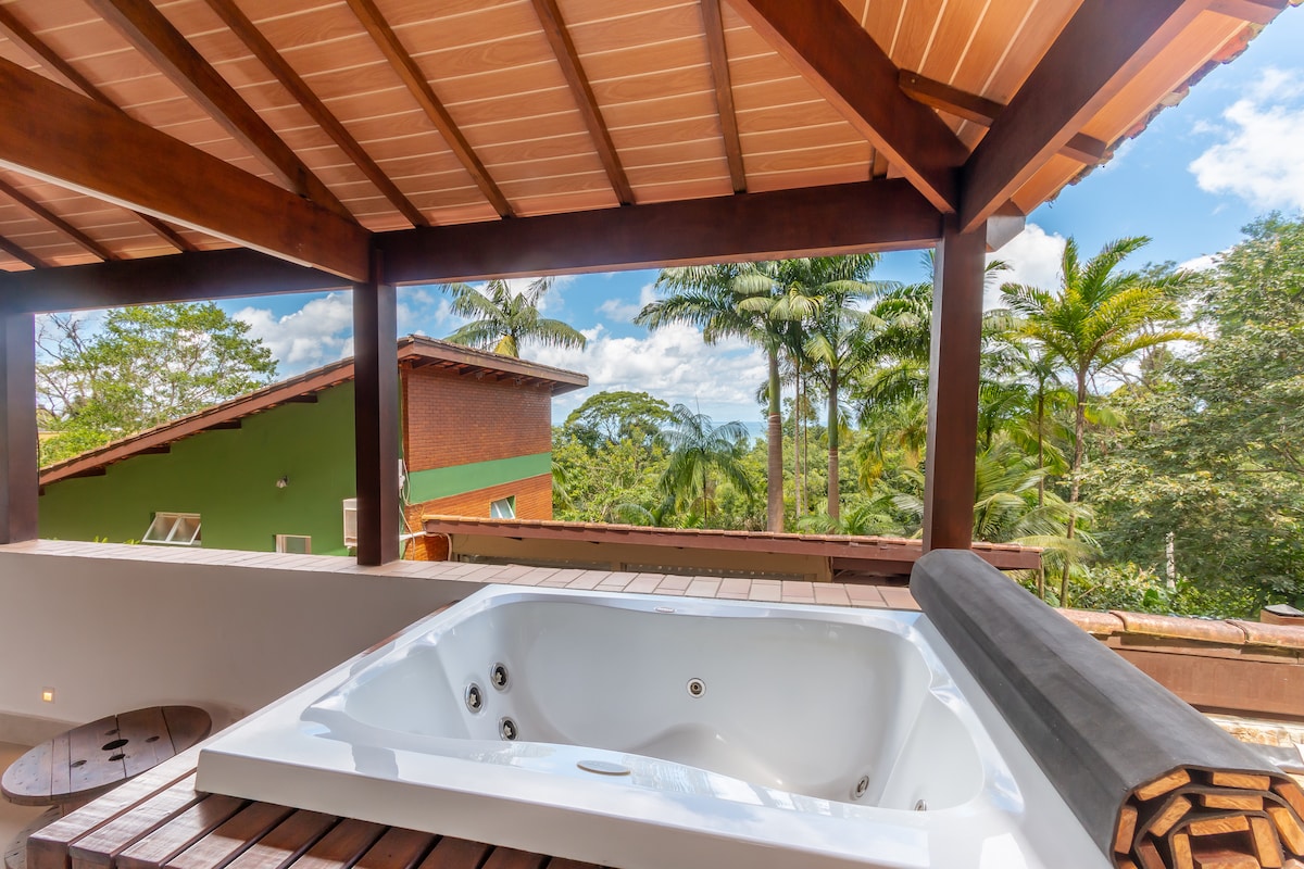 A jacuzzi is situated under a wooden pergola, surrounded by lush greenery and palm trees. The clear sky provides a bright backdrop, enhancing the serene atmosphere of the outdoor space.