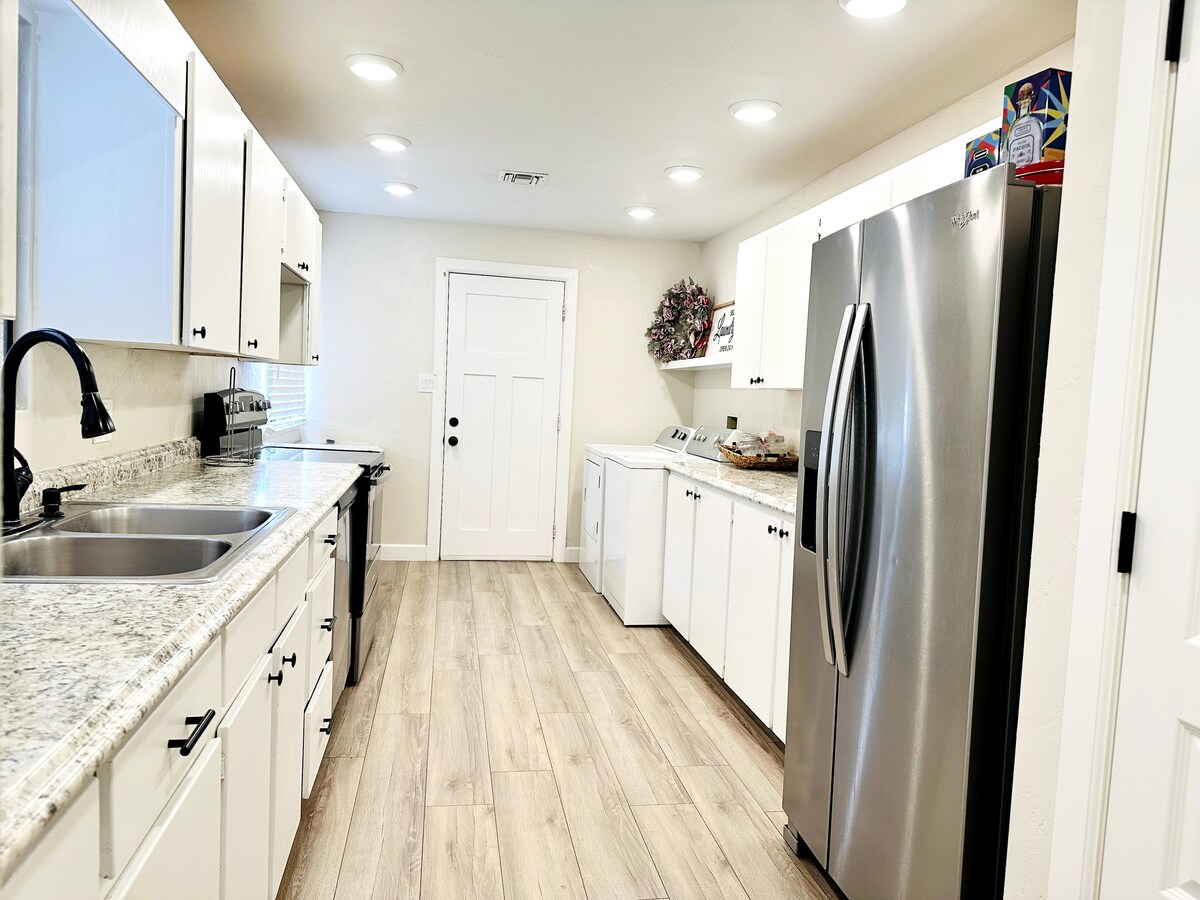 A modern kitchen is presented with white cabinetry and granite countertops. Stainless steel appliances, including a refrigerator and oven, enhance the space. Soft lighting from recessed fixtures brightens the area, while a door leads to the outside, providing easy access.