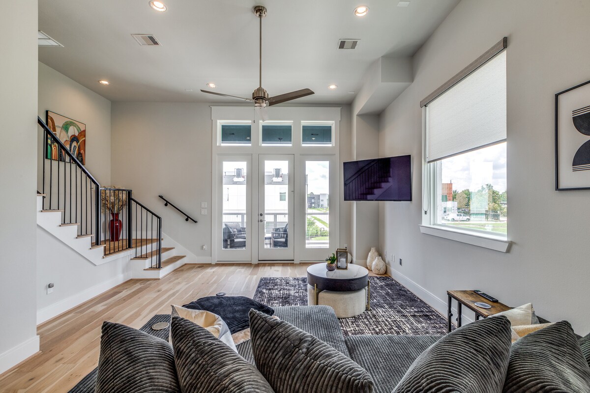 The modern living area features large windows allowing natural light to fill the space. A comfortable sofa is positioned near a round coffee table. A stylish rug adds texture to the floor, while a flat-screen TV is mounted on the wall, enhancing the inviting atmosphere.