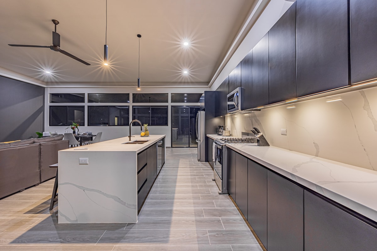 The spacious kitchen features modern, sleek cabinetry and a large island with a built-in sink. High-end appliances are arranged along the walls, while large glass windows allow natural light to fill the area. The open layout connects seamlessly with the dining space.