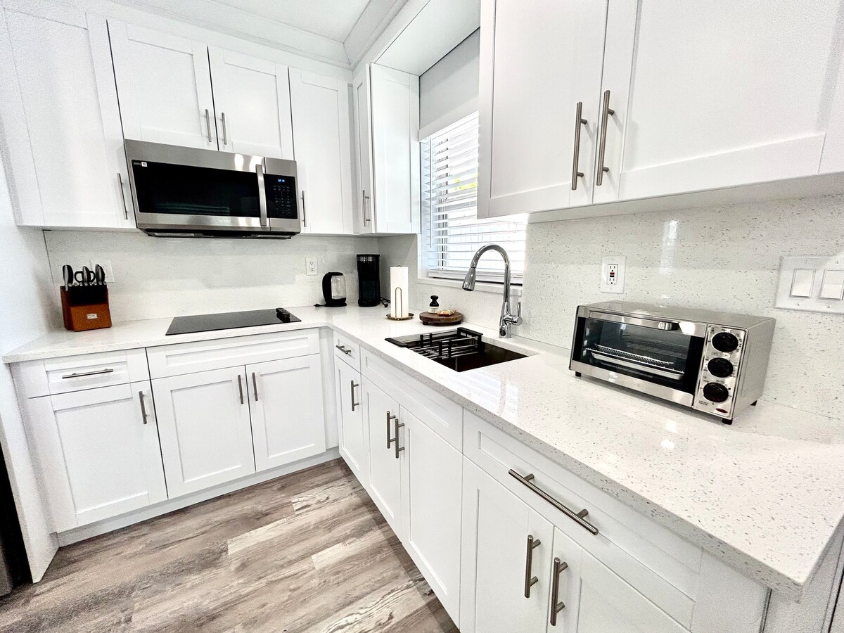 The well-equipped kitchen features modern white cabinets and sleek countertops. Stainless steel appliances, including a microwave and toaster oven, complement the space. Ample natural light filters in through a window, creating a bright atmosphere for cooking and meal preparation.