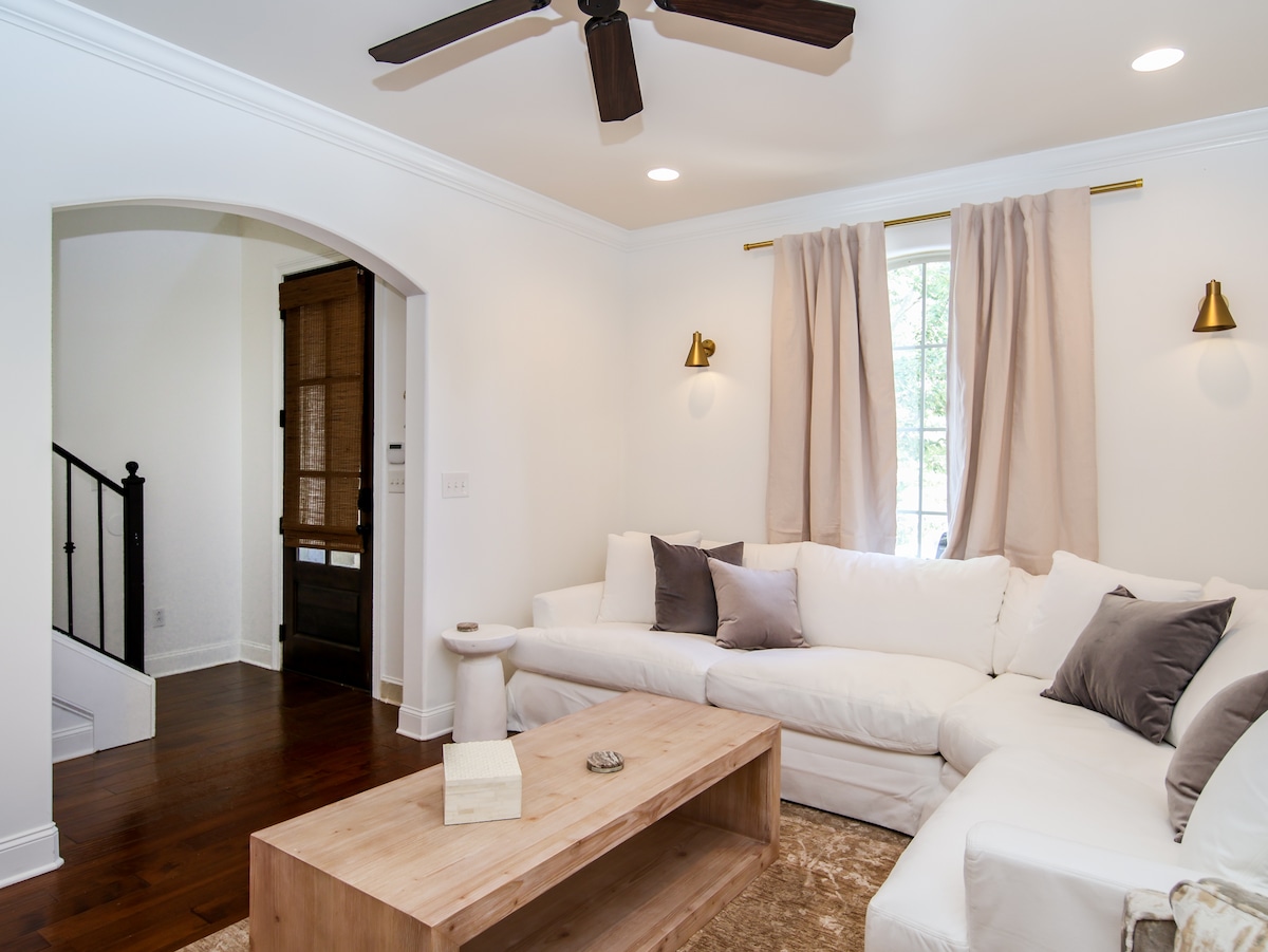 A light and airy living area features a spacious sectional sofa adorned with neutral-toned cushions. A simple wooden coffee table is positioned in front, complemented by soft, warm lighting from wall sconces. A window with flowing curtains provides natural light, enhancing the inviting ambience.