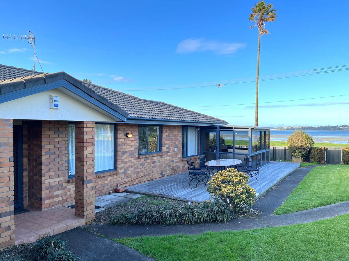 A single-story brick house features a patio area with a round dining table and chairs, surrounded by green grass and shrubs. Beyond, a scenic view of calm waters is visible, complemented by clear blue skies and a tall palm tree.