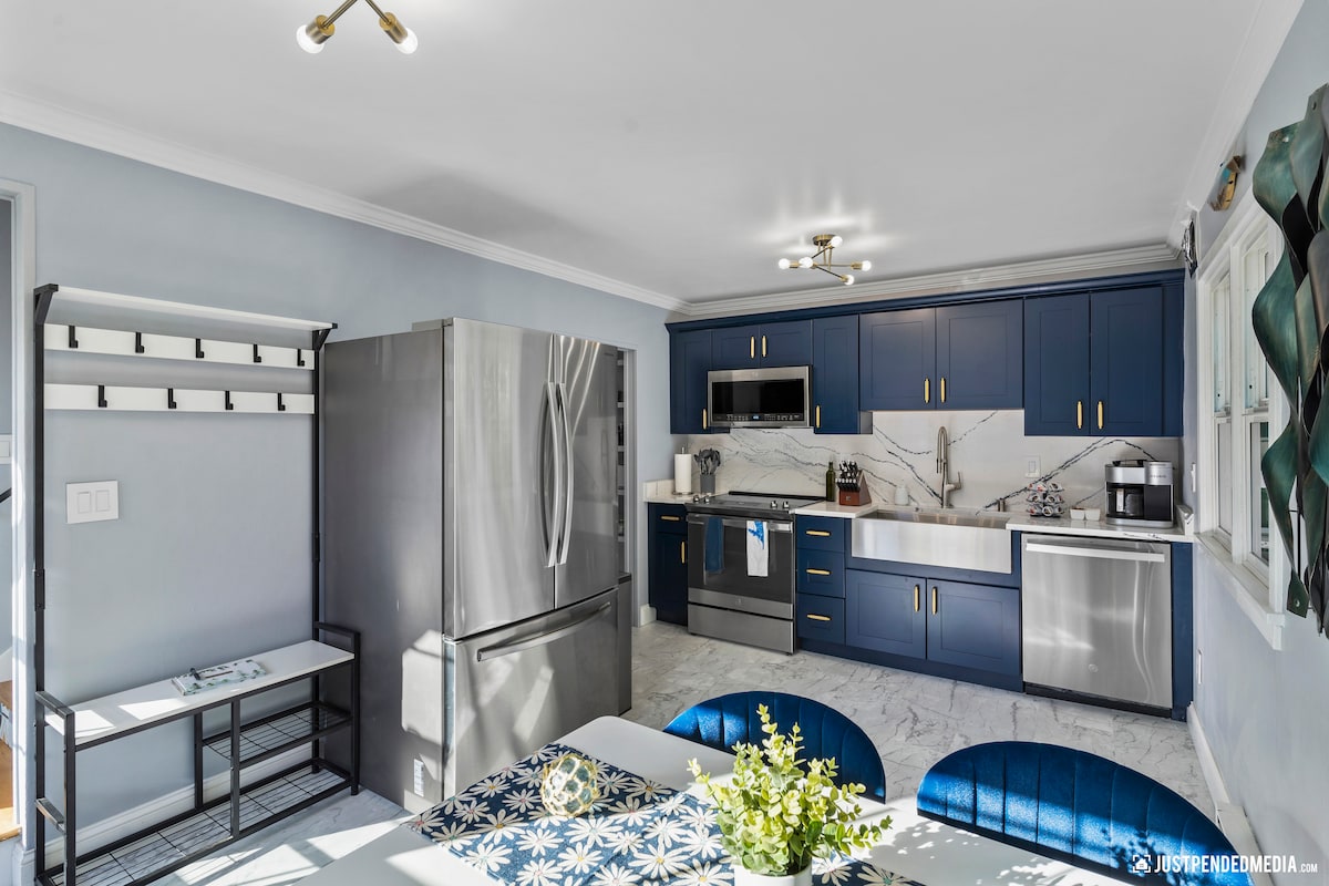 The kitchen features modern stainless steel appliances and a custom quartz countertop. A dining table with a decorative centerpiece is visible, complemented by velvet chairs. A storage bench and a wall-mounted rack provide convenient organization options. Natural light fills the space through the windows.