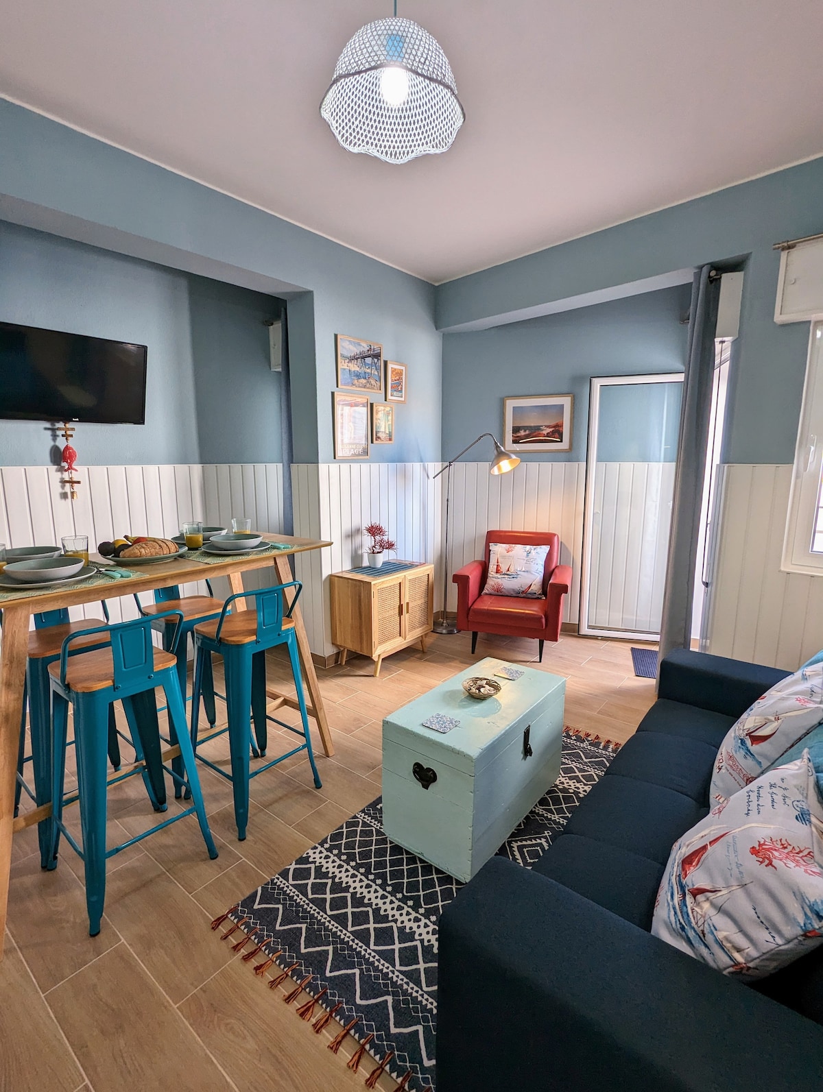 The living area features a cozy seating arrangement with a navy blue sofa and a red armchair. A small dining table is accompanied by turquoise bar stools. Natural light enters through a nearby door leading to the balcony, while coastal-themed decor adds to the vintage ambiance.