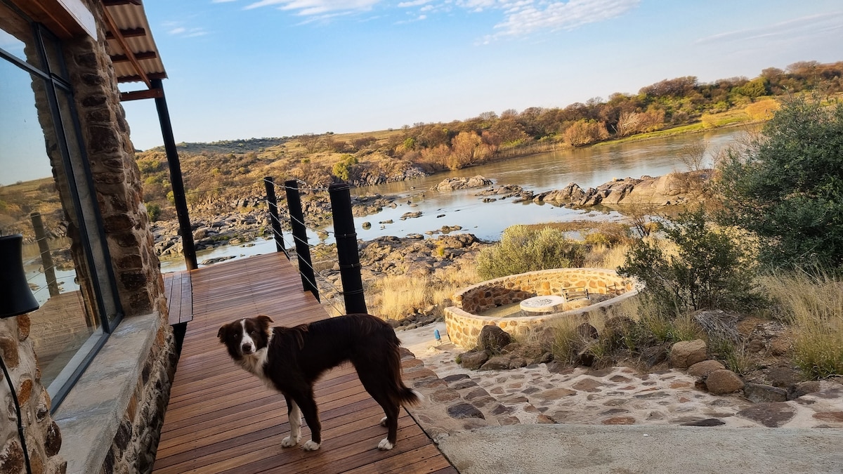 A wooden walkway leads towards the edge of a riverbank, with a dog standing nearby. The landscape includes rocky terrain and sparse vegetation, under a clear blue sky with soft clouds. A circular stone fire pit is visible in the foreground, situated on a grassy area.