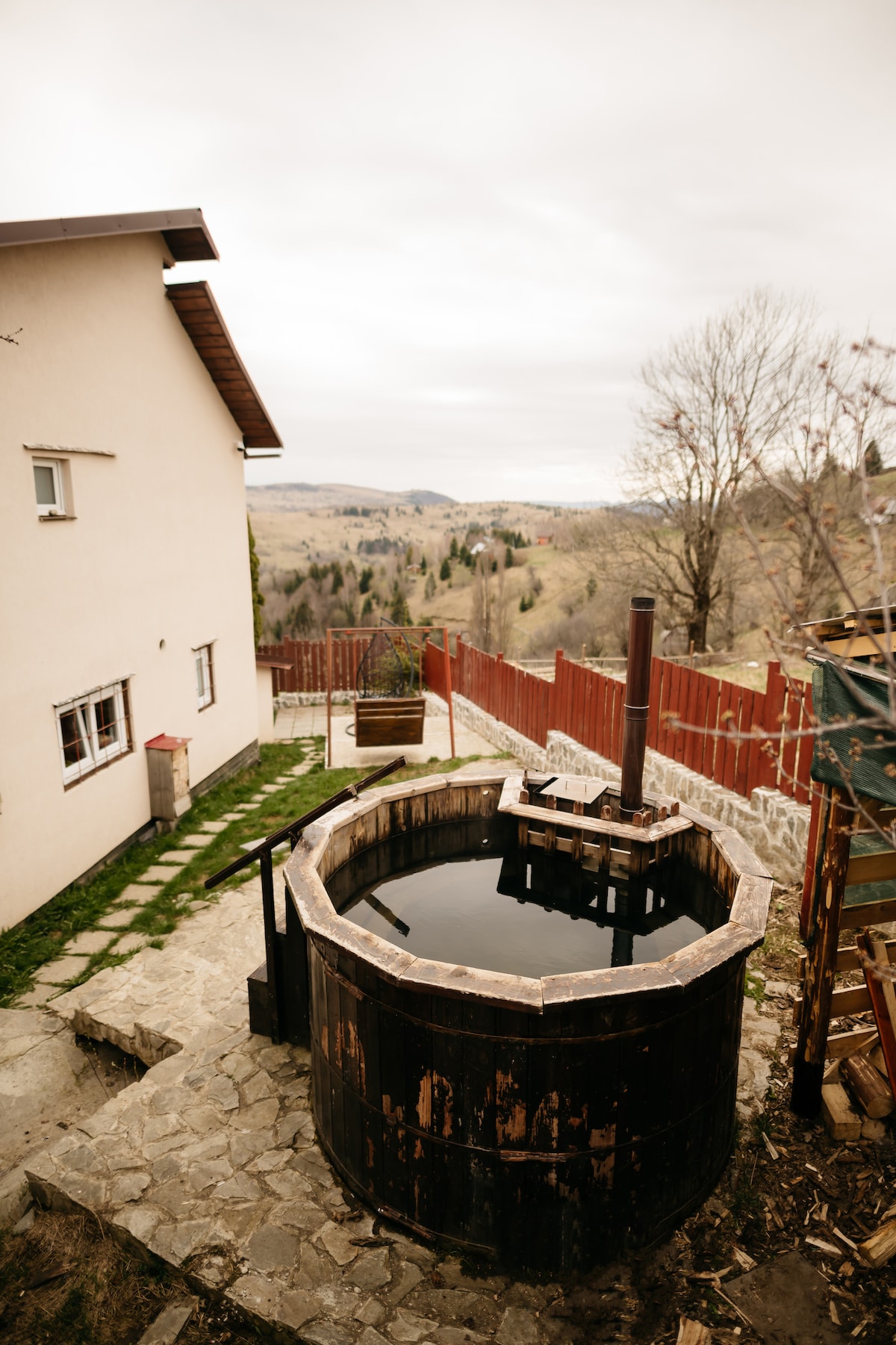 An outdoor space features a traditional wooden hot tub surrounded by natural stone. The view showcases rolling hills and trees in the distance, while a modern structure with a sloping roof is visible nearby. The area is enclosed by a wooden fence.