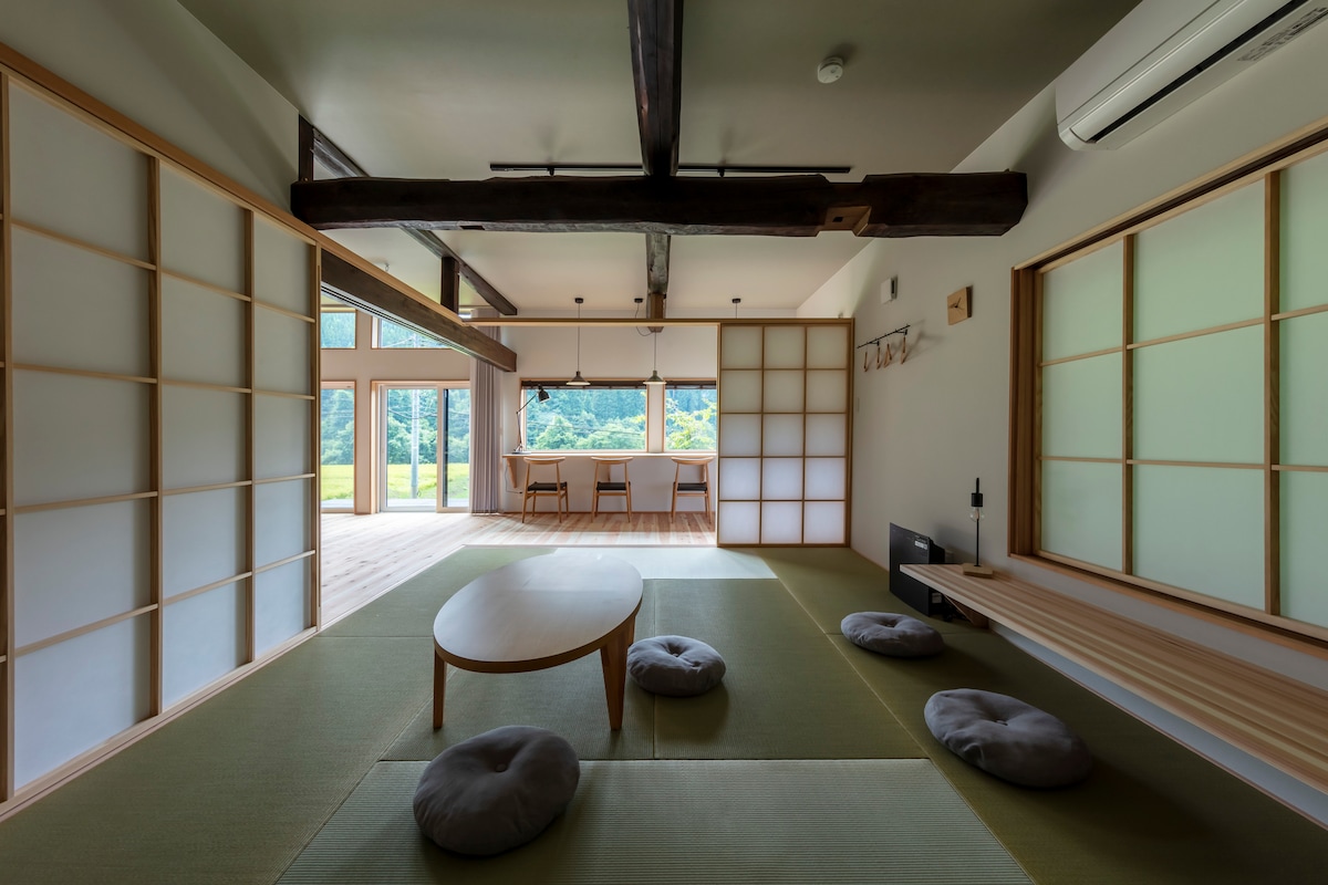 An inviting living space features tatami flooring and sliding shoji screens, allowing natural light to fill the room. A low wooden table is positioned on the floor, surrounded by round cushions. The view of the outdoors is enhanced by large glass doors that open to a serene landscape.