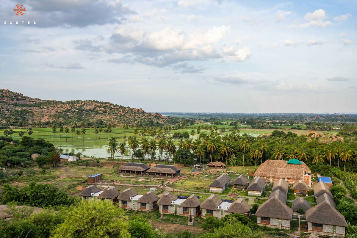 The image presents a scenic view of rustic hut-style accommodations surrounded by lush greenery. Palm trees and expansive fields are visible in the background, with gently rolling hills adding to the natural setting. A large building with a thatched roof stands prominently in the center.