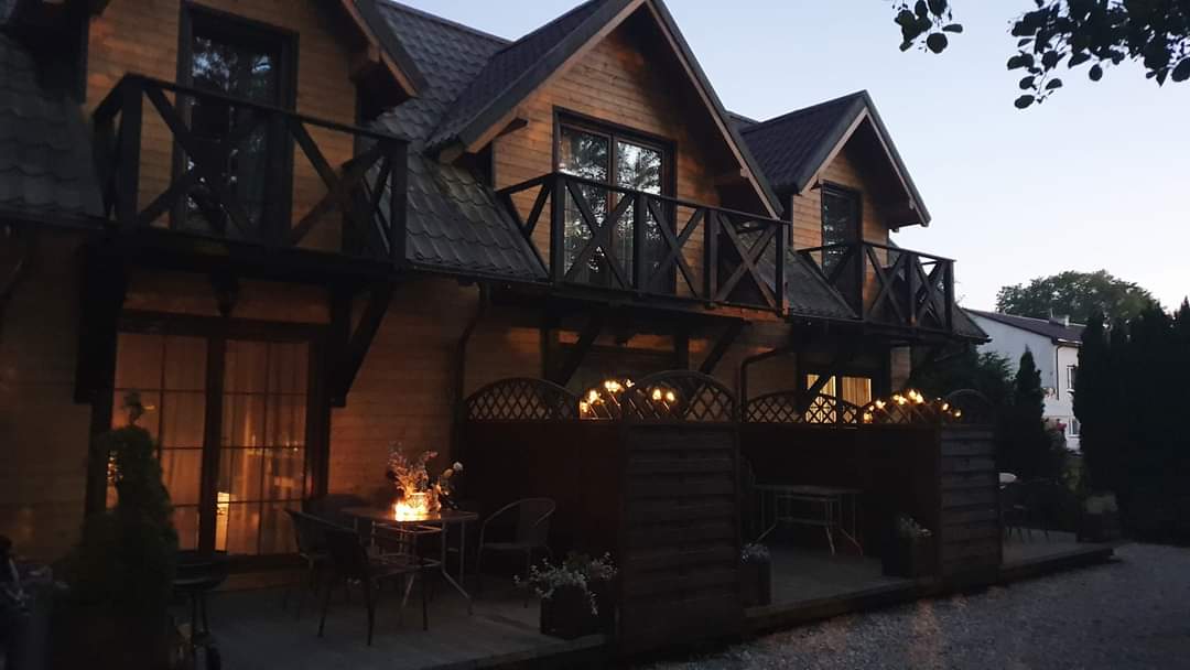 A row of wooden cottages is featured, each with a private balcony decorated with potted plants. Warm lighting illuminates the outdoor seating area, creating a welcoming ambiance for evening gatherings. A gravel pathway leads to the cottages, surrounded by well-maintained shrubbery.