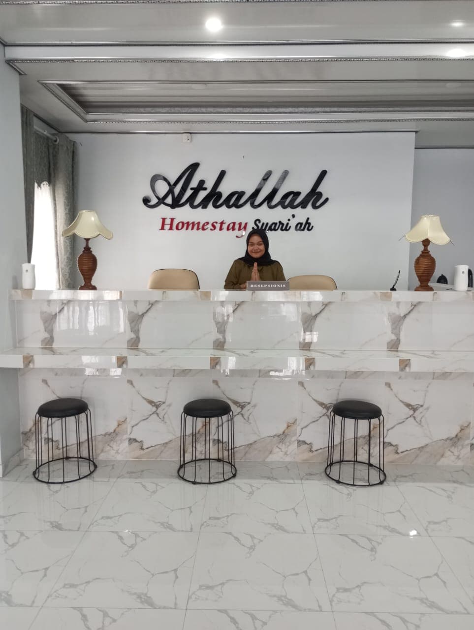The reception area features a sleek marble counter with three modern black stools arranged in front. A stylish light fixture is positioned on either side of the counter, creating a welcoming environment. The soft natural light enters through nearby windows.
