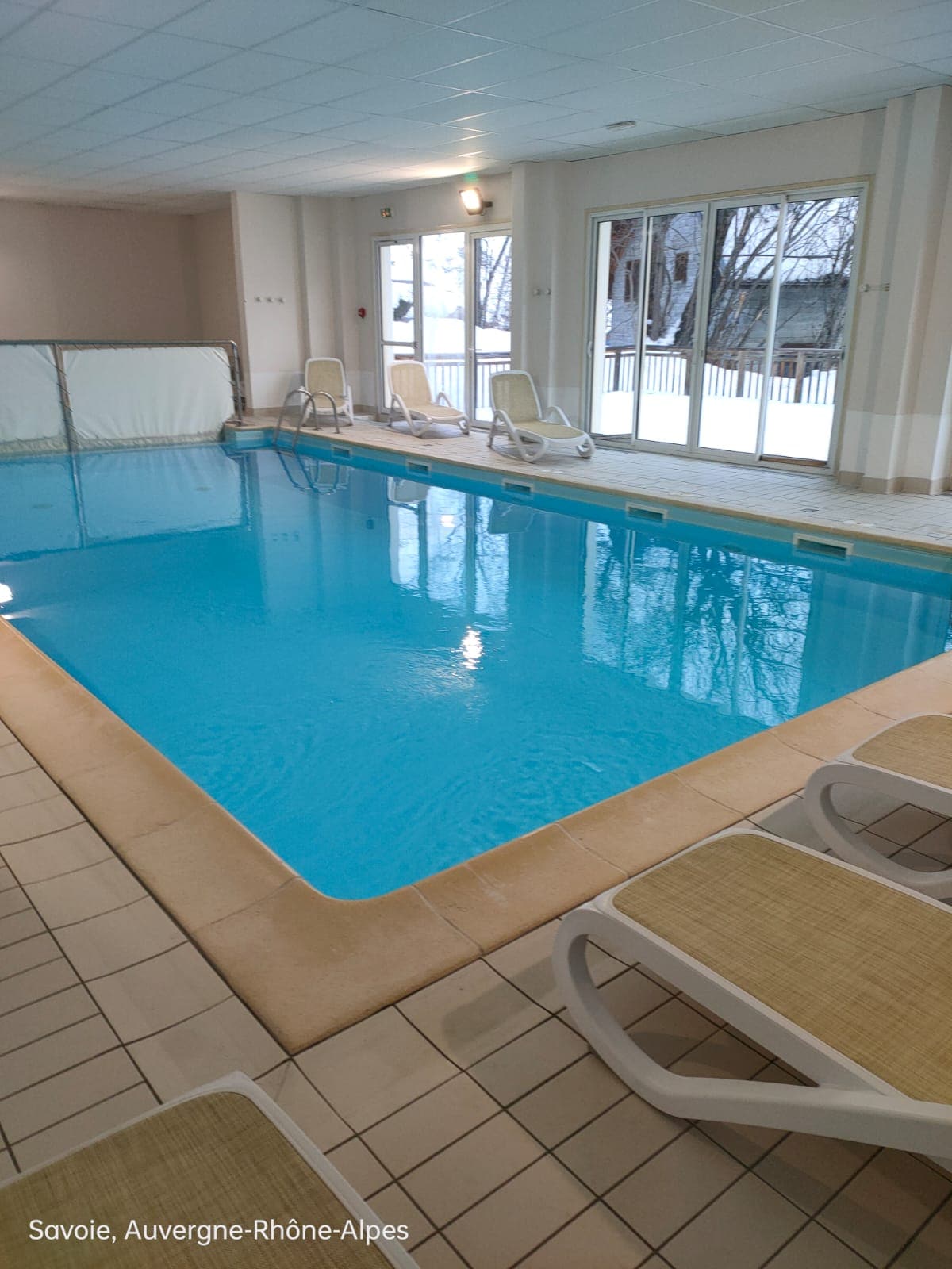 An indoor swimming pool is surrounded by large windows, allowing natural light to fill the space. Lounge chairs are placed along the poolside, and a protective cover is seen at one end of the pool. The area is designed for relaxation and enjoyment.