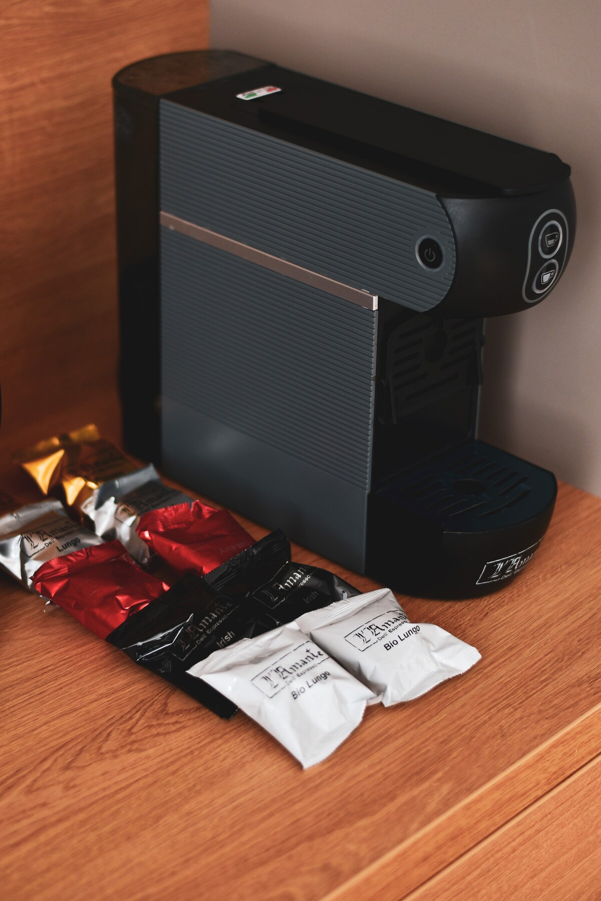 A coffee machine is placed on a wooden countertop, accompanied by several coffee pods in various colors, including red and silver. The machine is compact and designed for easy use, contributing to a convenient coffee-making experience.
