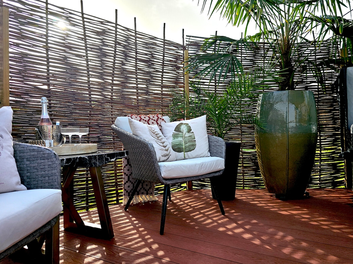 A private outdoor seating area features modern wicker chairs with plush cushions, complemented by a decorative table. Lush greenery and a large planter surround the space, while a sun-drenched atmosphere lends a relaxed ambiance.
